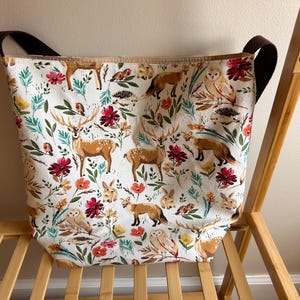 May include: A white canvas tote bag with brown straps features a colorful print of woodland animals, including deer, owls, and foxes. The print also includes flowers and leaves.