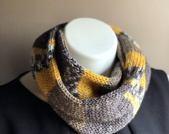 May include: A knitted infinity scarf in shades of gray, brown, and yellow. The scarf is displayed on a white mannequin head, showcasing its geometric color block pattern. The soft texture and warm tones suggest a cozy accessory.