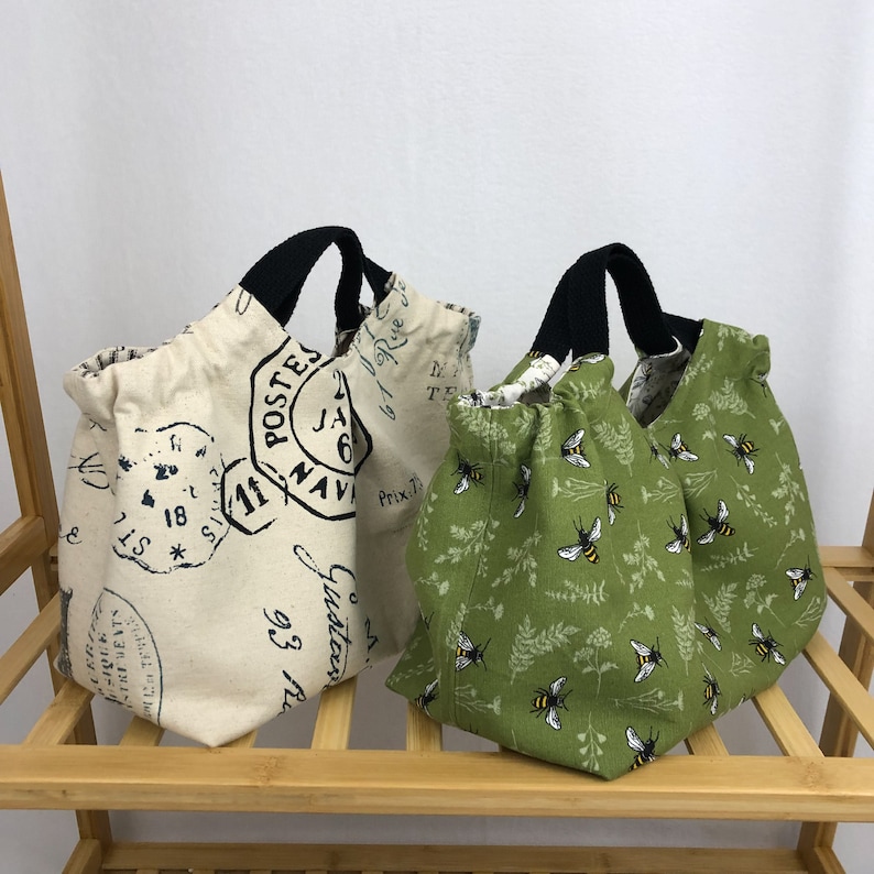 May include: Two reusable shopping bags. One is beige with black straps and a vintage French postage stamp design. The other is green with black straps and a bee pattern.