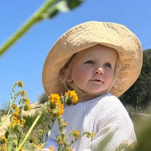 May include: A young child wearing a white sweater and a light brown sun hat is standing in a field of yellow wildflowers. The child is looking to the right of the frame.