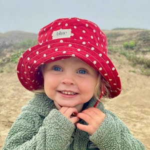 May include: A young child wearing a red and white polka dot bucket hat with the word "blue" on the front. The child is wearing a green fleece jacket and is smiling at the camera.