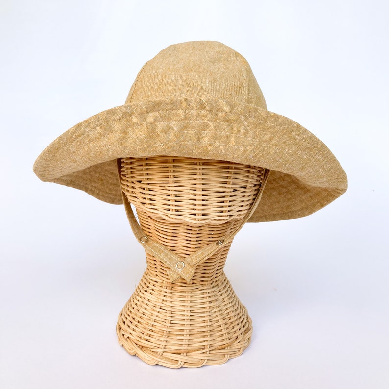 Wide brim tan colored linen/cotton hand made sun hat on rattan baby head mannequin. Chin straps close with two snaps for adustable fit. Brim can flip up or stay down with ease.