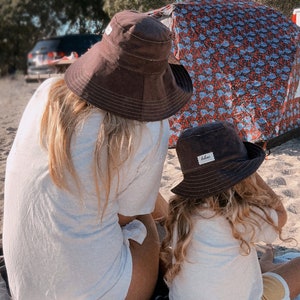 May include: Two people wearing brown and black bucket hats are sitting on a beach blanket. The person in the foreground is wearing a white t-shirt and has long blonde hair. The person in the background is wearing a white t-shirt and has long brown hair. The hat in the foreground has a small white label that says "blue".