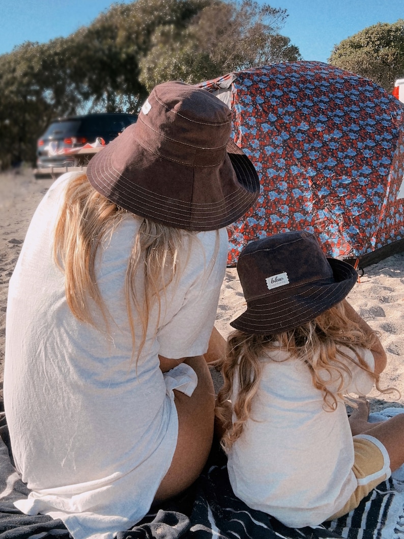 May include: Two people wearing brown and black bucket hats are sitting on a beach blanket. The person in the foreground is wearing a white t-shirt and has long blonde hair. The person in the background is wearing a white t-shirt and has long brown hair. The hat in the foreground has a small white label that says "blue".