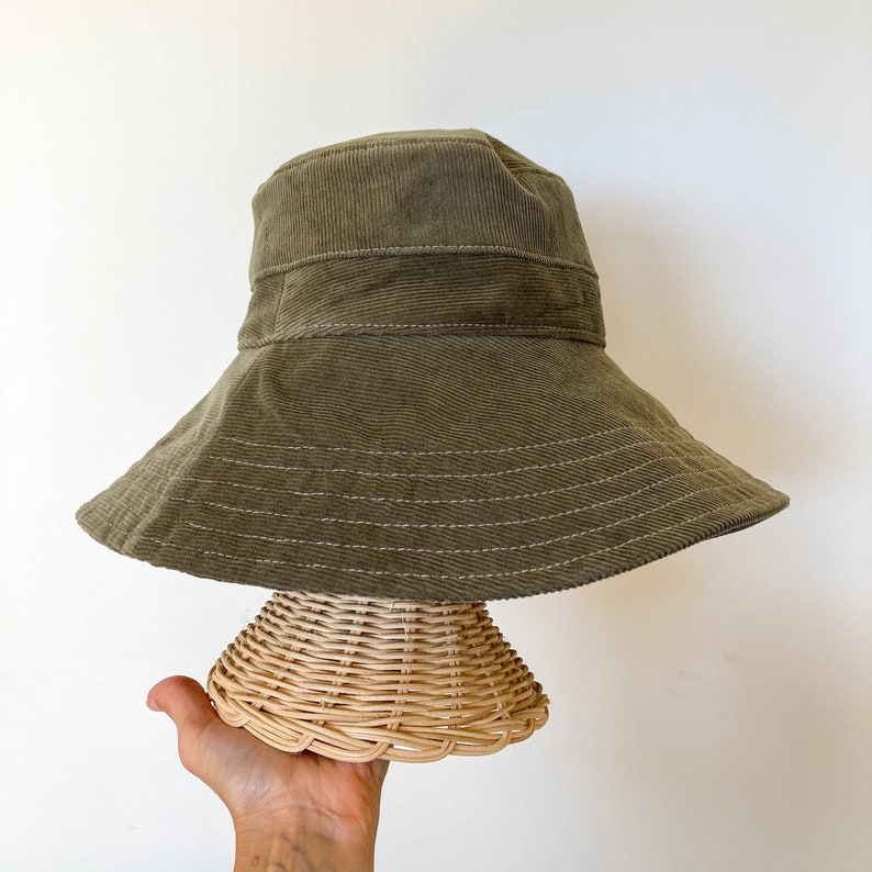 K&ouml;nnte beinhalten: Ein breiter gr&uuml;ner Cord-Bucket-Hut mit einem gesteppten Design. Der Hut wird auf einer Hutform aus Weidengeflecht pr&auml;sentiert.