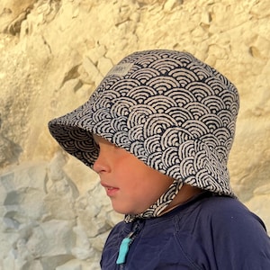May include: A navy and white patterned sun hat with a wide brim. The hat features a repeating wave-like design and a small label. The hat has chin straps and is worn by a person.