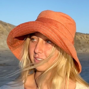May include: A wide-brimmed, orange corduroy bucket hat with white stitching detail. The hat is being worn by a person with blonde hair.