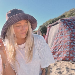May include: A woman wearing a white t-shirt and a dark grey bucket hat sits on a sandy beach. She has long blonde hair and is wearing a necklace. A colourful tent is in the background.