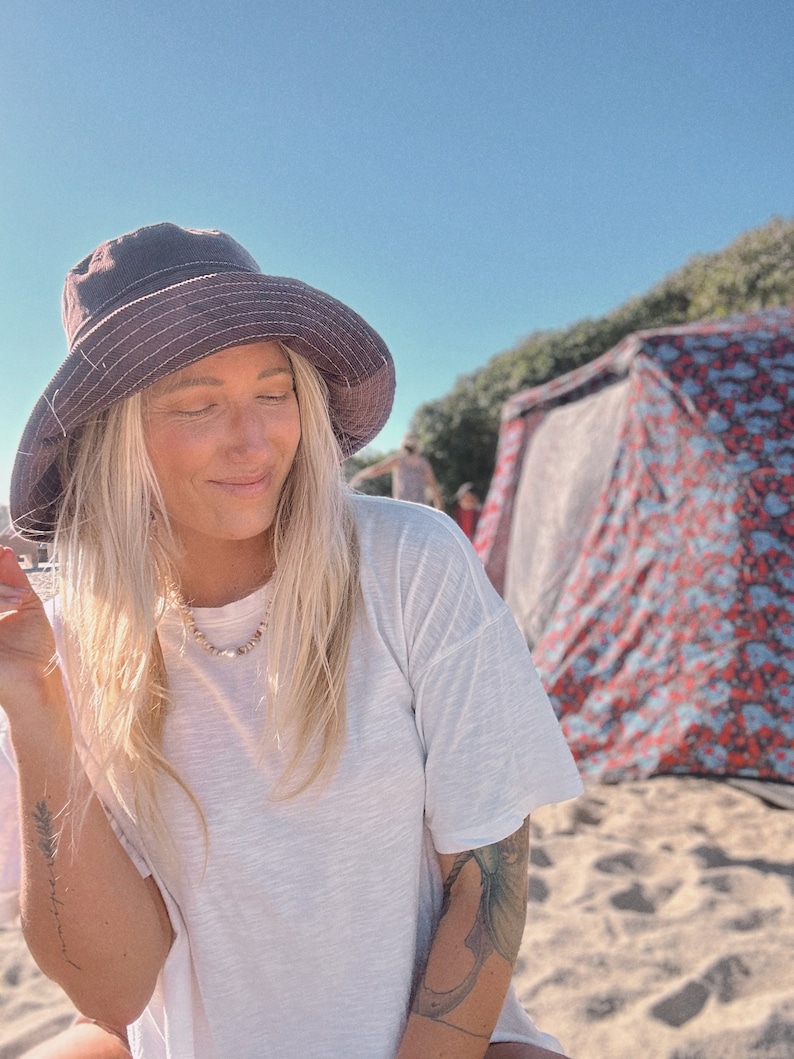 May include: A woman wearing a white t-shirt and a dark grey bucket hat sits on a sandy beach. She has long blonde hair and is wearing a necklace. A colourful tent is in the background.