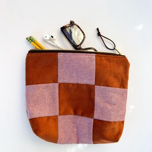 May include: A brown and pink checkered corduroy zippered pouch with a brown zipper. The pouch is open, revealing two pencils, white earbuds, and eyeglasses. The eyeglasses have a tortoiseshell pattern.