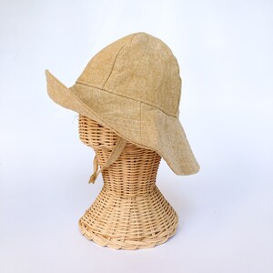 May include: A wide-brimmed sun hat made of a light brown linen fabric. The hat is sitting on a wicker hat stand.