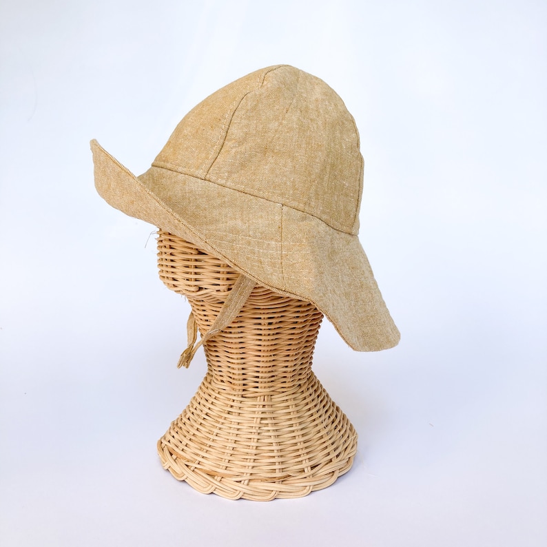 May include: A wide-brimmed sun hat made of a light brown linen fabric. The hat is sitting on a wicker hat stand.