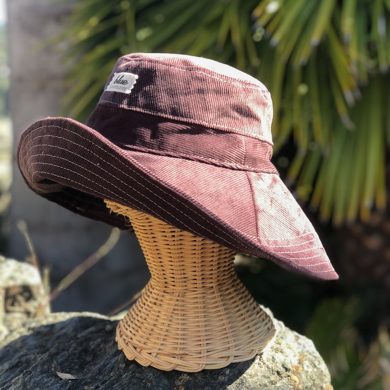 May include: A wide-brimmed hat made of brown corduroy with a white label that reads "Blue Cotton". The hat is displayed on a wicker hat stand.