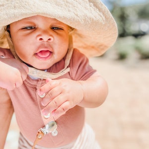 May include: A baby wearing a light brown sun hat, a pink ribbed onesie, and white and brown striped trousers. The baby is looking at the camera and has its tongue sticking out.