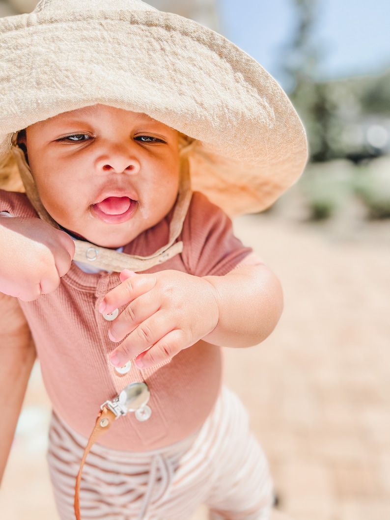May include: A baby wearing a light brown sun hat, a pink ribbed onesie, and white and brown striped trousers. The baby is looking at the camera and has its tongue sticking out.