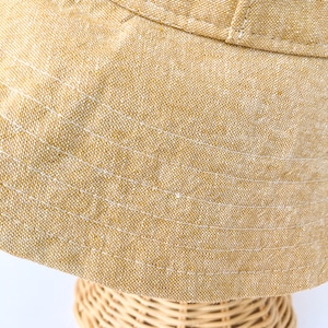 May include: A close-up of a light brown bucket hat made of a textured fabric with white stitching. The hat is folded down and has a wide brim.