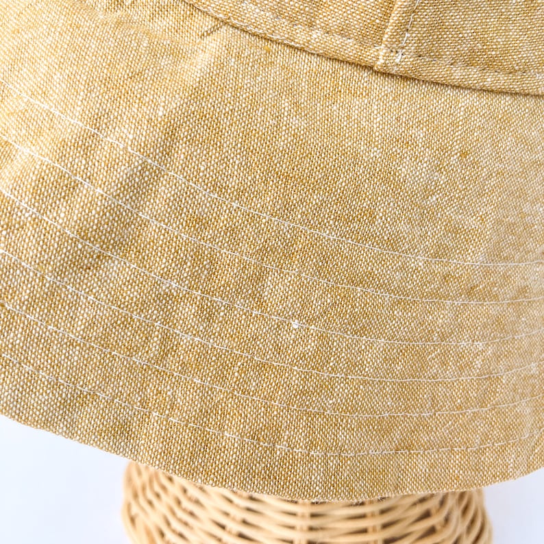 May include: A close-up of a light brown bucket hat made of a textured fabric with white stitching. The hat is folded down and has a wide brim.