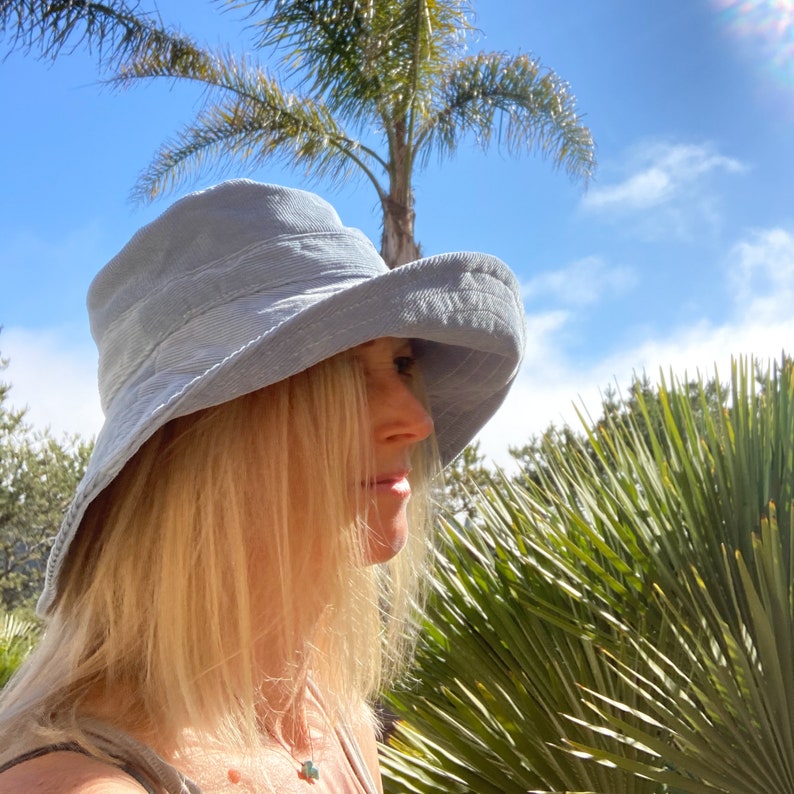 Pu&ograve; includere: Un cappello da sole a tesa larga di colore azzurro chiaro con un tessuto leggermente strutturato. Il cappello &egrave; indossato da una persona con i capelli lunghi e biondi.