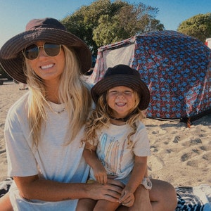 May include: A woman wearing a brown hat and sunglasses is holding a young girl wearing a brown hat and a white t-shirt with a colourful surfboard design. The girl is sitting on the woman's lap. They are sitting on a beach with a tent in the background.