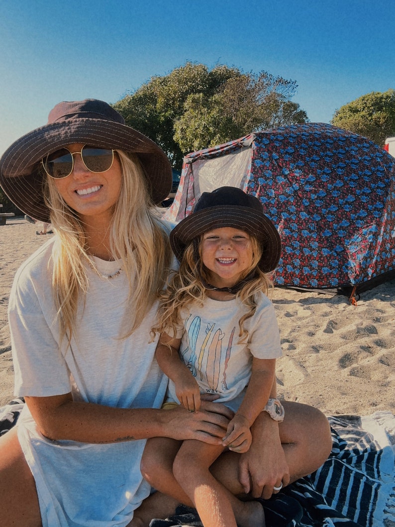 May include: A woman wearing a brown hat and sunglasses is holding a young girl wearing a brown hat and a white t-shirt with a colourful surfboard design. The girl is sitting on the woman's lap. They are sitting on a beach with a tent in the background.