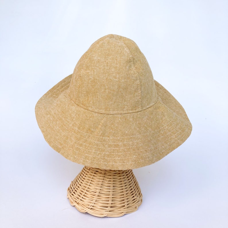 May include: A tan linen sun hat with a wide brim and a rounded crown. The hat is displayed on a woven wicker hat stand.