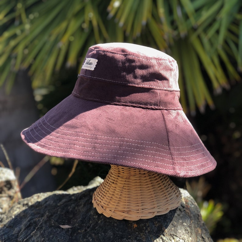 May include: A wide-brimmed sun hat made of brown corduroy with a white label that says "Blue " on it. The hat is sitting on a wicker hat stand.
