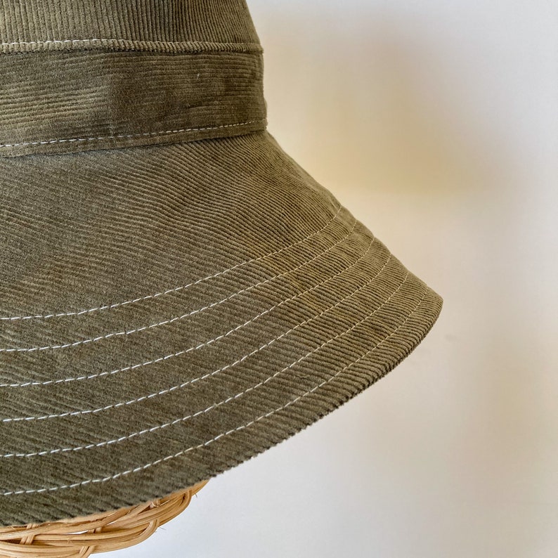 K&ouml;nnte beinhalten: Ein gr&uuml;ner Cord-Bucket-Hut mit wei&szlig;en N&auml;hten. Der Hut hat eine breite Krempe und eine flache Krone.