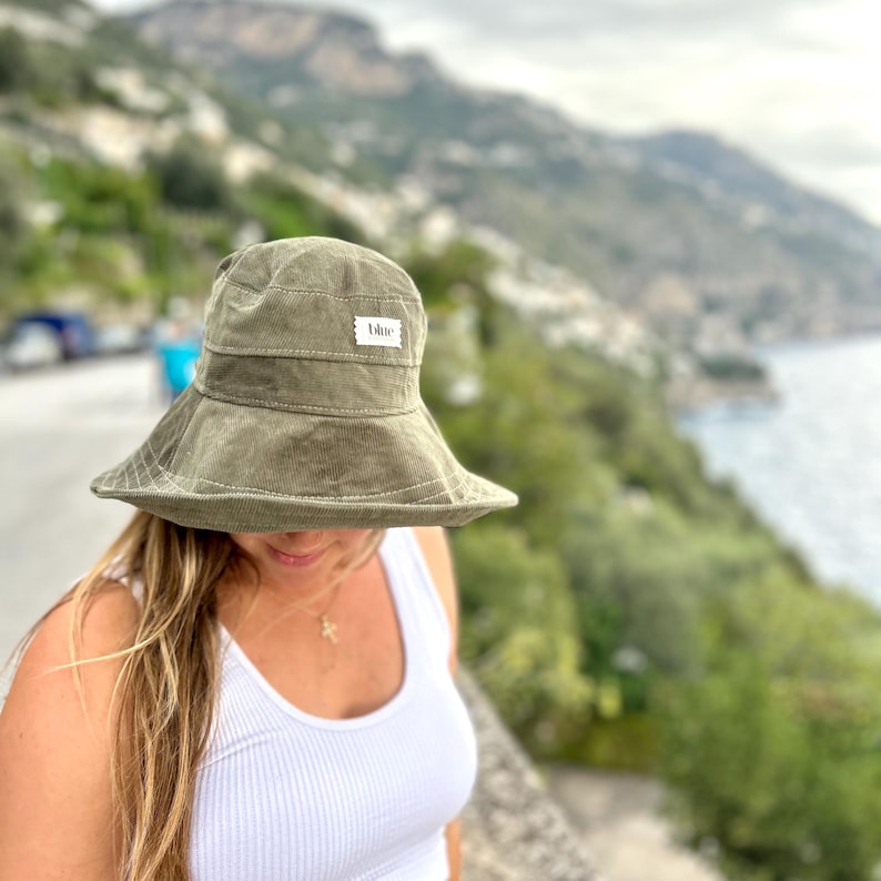 K&ouml;nnte beinhalten: Eine Person tr&auml;gt einen gr&uuml;nen Cord-Bucket-Hut mit einem wei&szlig;en Etikett, auf dem "Ulule" steht. Der Hut wird &uuml;ber dem Haar der Person getragen und bedeckt ihre Stirn.