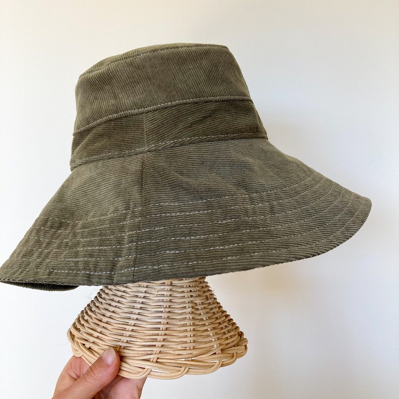 K&ouml;nnte beinhalten: Ein breiter, olivgr&uuml;ner Cord-Bucket-Hut mit einer darunterliegenden Form aus geflochtenem Stroh.