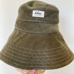 K&ouml;nnte beinhalten: Ein gr&uuml;ner Cord-Bucket-Hut mit einem wei&szlig;en Etikett, auf dem "blue corduroy" steht.