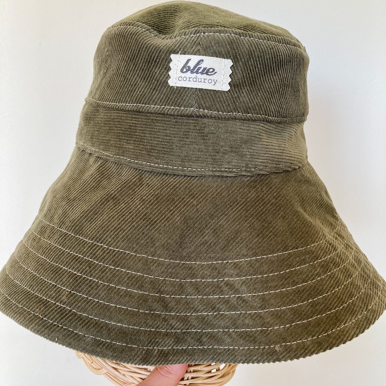 K&ouml;nnte beinhalten: Ein gr&uuml;ner Cord-Bucket-Hut mit einem wei&szlig;en Etikett, auf dem "blue corduroy" steht.