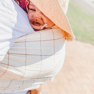 May include: A baby wearing a beige and white checked wrap carrier and a tan sun hat with a dummy in their mouth.