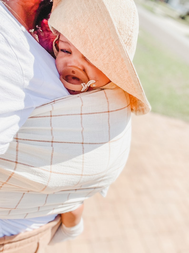May include: A baby wearing a beige and white checked wrap carrier and a tan sun hat with a dummy in their mouth.