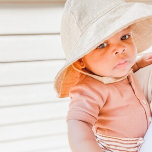 May include: A baby wearing a light brown sun hat and a peach coloured ribbed onesie with brown and white stripes. The baby is looking at the camera.