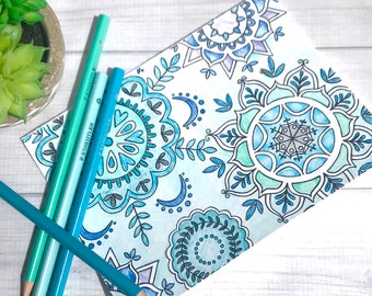 May include: A close-up of a colouring page with intricate blue and green mandala designs. The page is lying on a white wooden surface with several blue coloured pencils beside it.