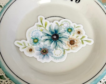 May include: A floral sticker with a watercolour design of blue, teal, and cream flowers with green leaves. The sticker is on a white plate with a blue rim.