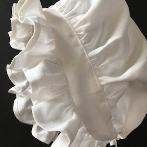 May include: A white linen bonnet with ruffled trim and ties. The word "Linen" is printed in white at the bottom of the image. The bonnet is made of a soft, lightweight fabric.
