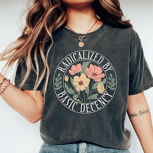 Peut inclure: T-shirt gris foncé avec un motif floral à l'intérieur d'un cercle blanc. Le texte indique "RADICALIZED BY BASIC DECENCY" en cercle autour des fleurs. Les fleurs sont roses et jaunes avec des feuilles vertes.
