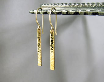 May include: A pair of gold hammered bar earrings. The earrings are long and thin, with a hammered texture. They are hanging from a silver stand.