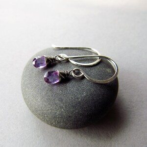 K&ouml;nnte beinhalten: Ein Paar silberne Ohrringe mit kleinen violetten Amethyststeinen. Die Ohrringe h&auml;ngen an einem grauen Stein.