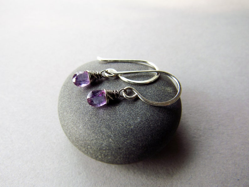 K&ouml;nnte beinhalten: Ein Paar silberne Ohrringe mit kleinen violetten Amethyststeinen. Die Ohrringe h&auml;ngen an einem grauen Stein.