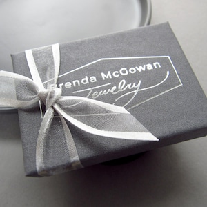 K&ouml;nnte beinhalten: Eine graue Schmuckschatulle mit einem wei&szlig;en Band und dem Aufdruck "Brenda McGowan Jewelry" auf der Oberseite.