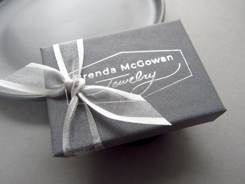 K&ouml;nnte beinhalten: Eine graue Schmuckschatulle mit einem wei&szlig;en Band und dem Aufdruck "Brenda McGowan Jewelry" auf der Oberseite.