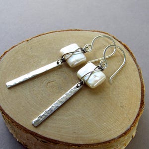 May include: A pair of handcrafted earrings featuring white, square-shaped pearls wrapped in wire, suspended from silver-toned hooks. The earrings have hammered, rectangular silver bars. The jewelry rests on a wooden surface.