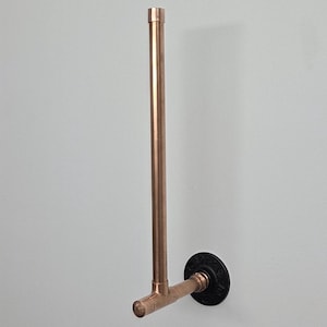 May include: A copper-colored pipe towel rack mounted on a white wall. The rack is made of copper pipes and fittings, with a black circular base. The design is industrial and minimalist.