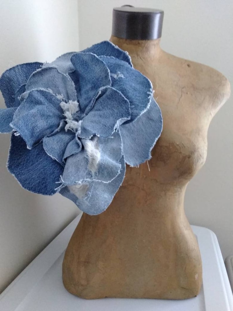 May include: A large denim flower, made from multiple layers of blue denim fabric, is displayed on a brown mannequin torso.