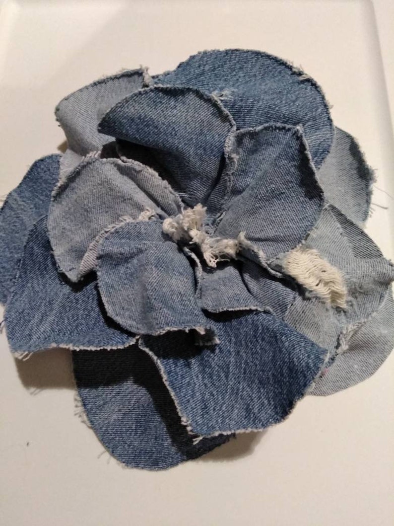 May include: A blue denim flower made from recycled fabric scraps. The flower is made up of several layers of denim, each with a slightly different shade of blue. The edges of the denim are frayed and uneven, giving the flower a rustic look.