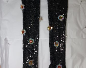 May include: A pair of long black sequined gloves with floral embellishments. The gloves have a sheer mesh fabric and are adorned with iridescent sequins and colorful flower-shaped appliques.