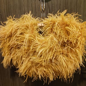 May include: A fluffy, golden-brown feather boa, hanging from a silver hanger. The boa is densely packed with soft, feathery plumes, creating a voluminous and textured appearance. The background is a dark, wood-paneled wall.