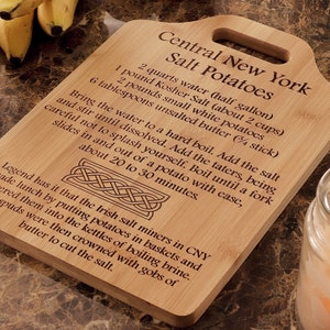 May include: A wooden cutting board with a recipe for Central New York Salt Potatoes. The recipe is engraved on the board and includes ingredients like water, salt, and potatoes. The board has a handle and a Celtic knot design.